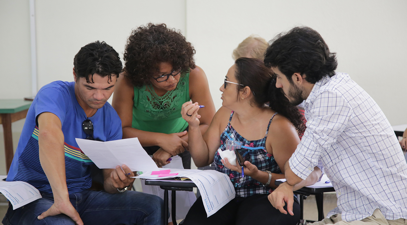 Retrato de quatro pessoas conversando, uma delas está lendo um texto em uma folha branca e os outros três estão gesticulando e debatendo sobre o tema.